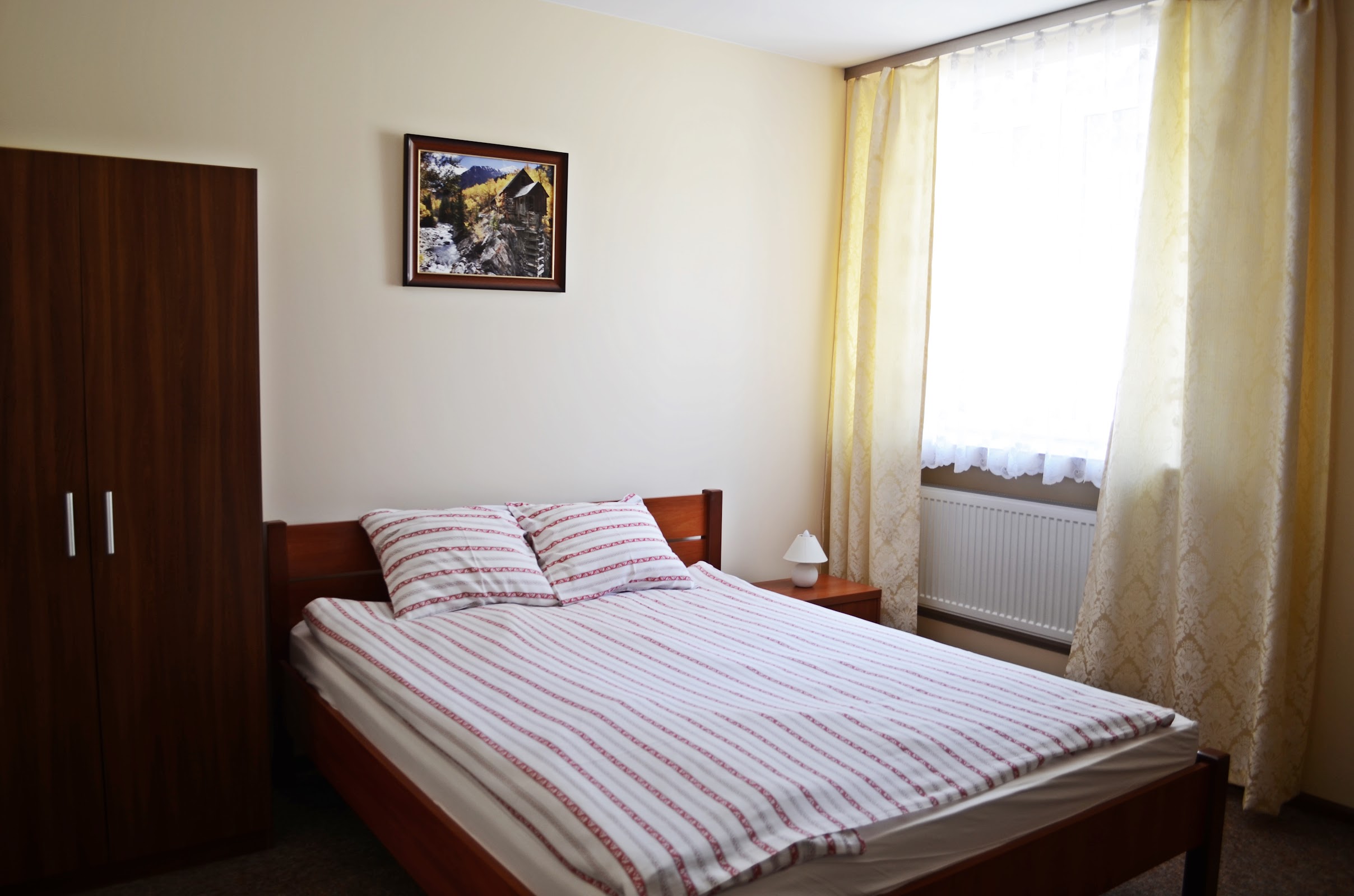 Pokój dwuosobowy w Willi Sailor - drewniane łóżko z biało-czerwoną pościelą, drewniana szafa, obraz nad łóżkiem, jasne ściany, żółte zasłony, kaloryfer pod oknem