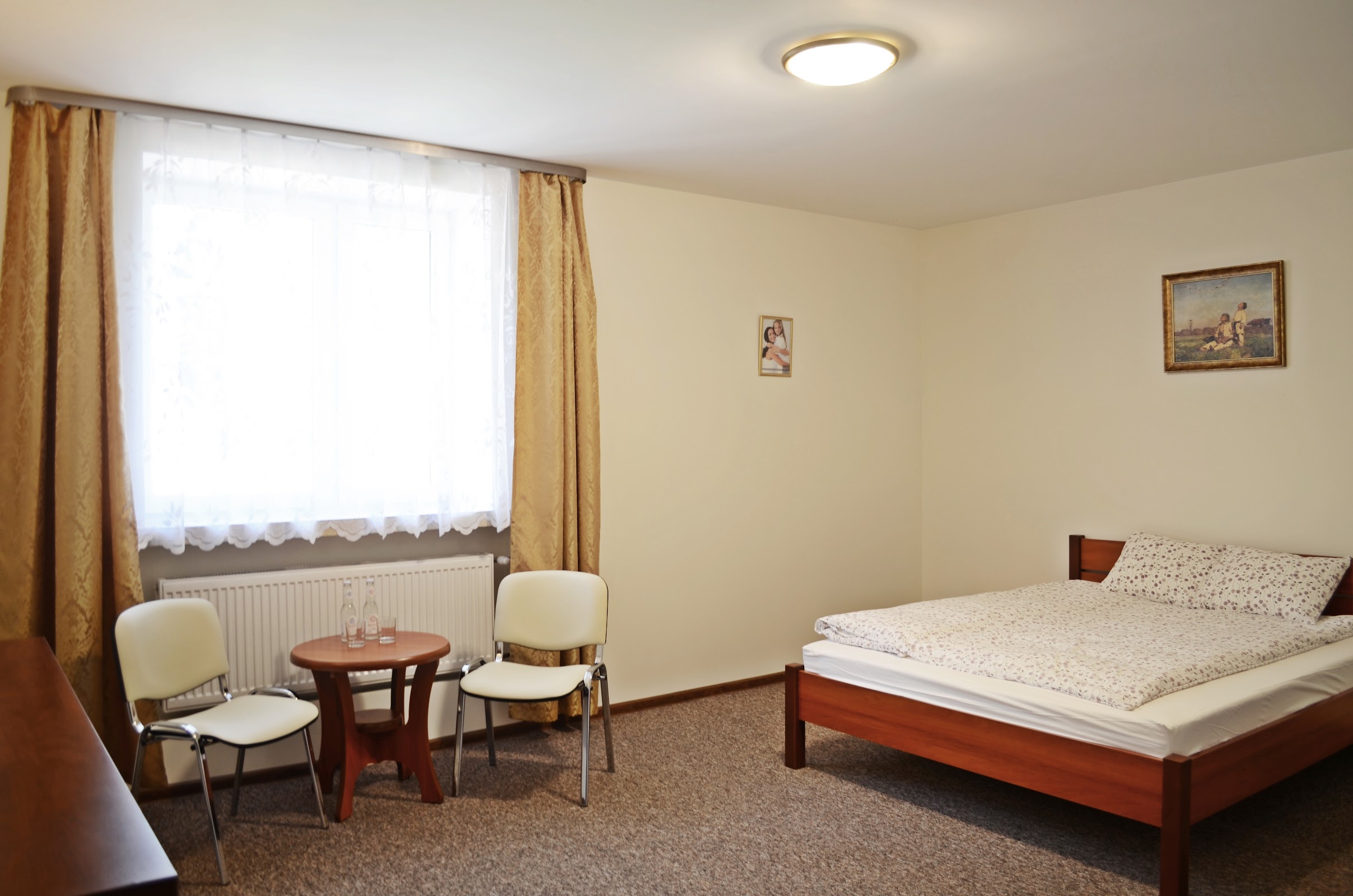 Pokój trzyosobowy w Willi Sailor - przestronne wnętrze, duże łóżko, dwa krzesła i okrągły stolik przy oknie, jasne beżowe ściany, drewniane meble, dwa obrazy na ścianie
