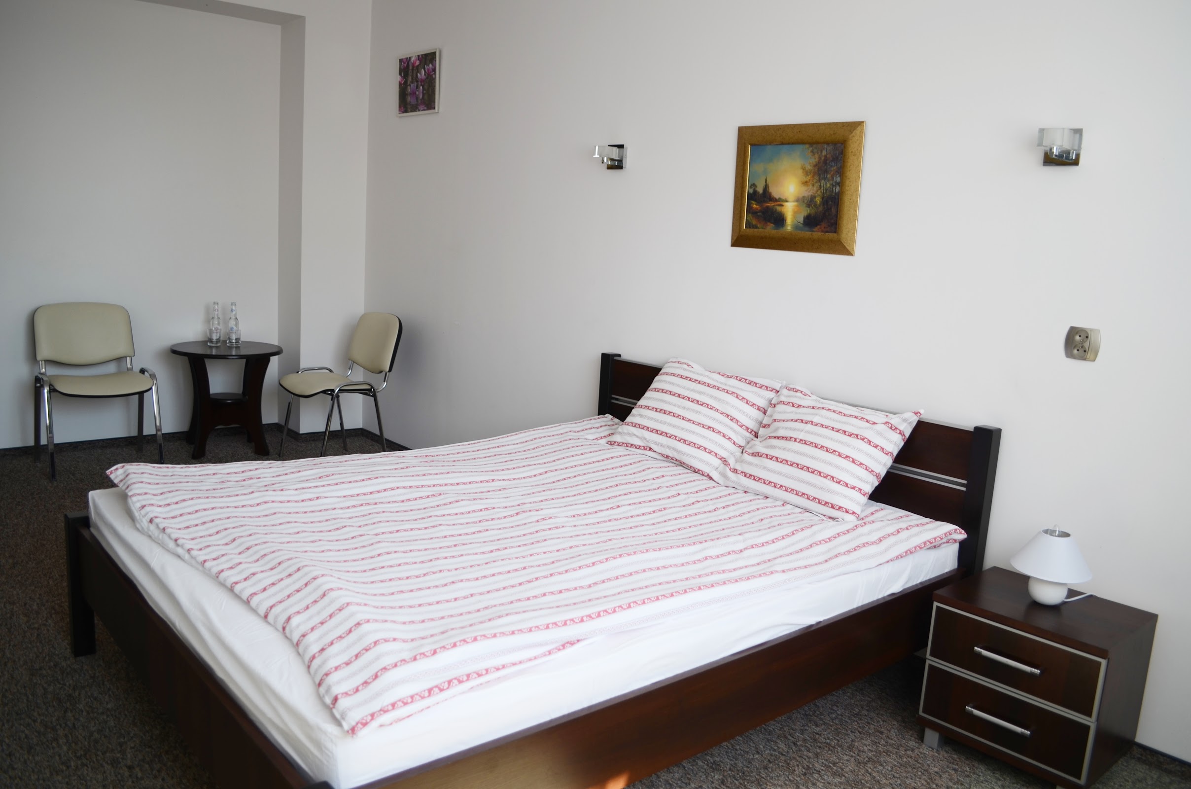 Apartament w Willi Sailor - duże łóżko małżeńskie z ciemną ramą, kącik wypoczynkowy z dwoma fotelami i stolikiem, obraz pejzażowy w złotej ramie, jasne ściany i drewniane akcenty